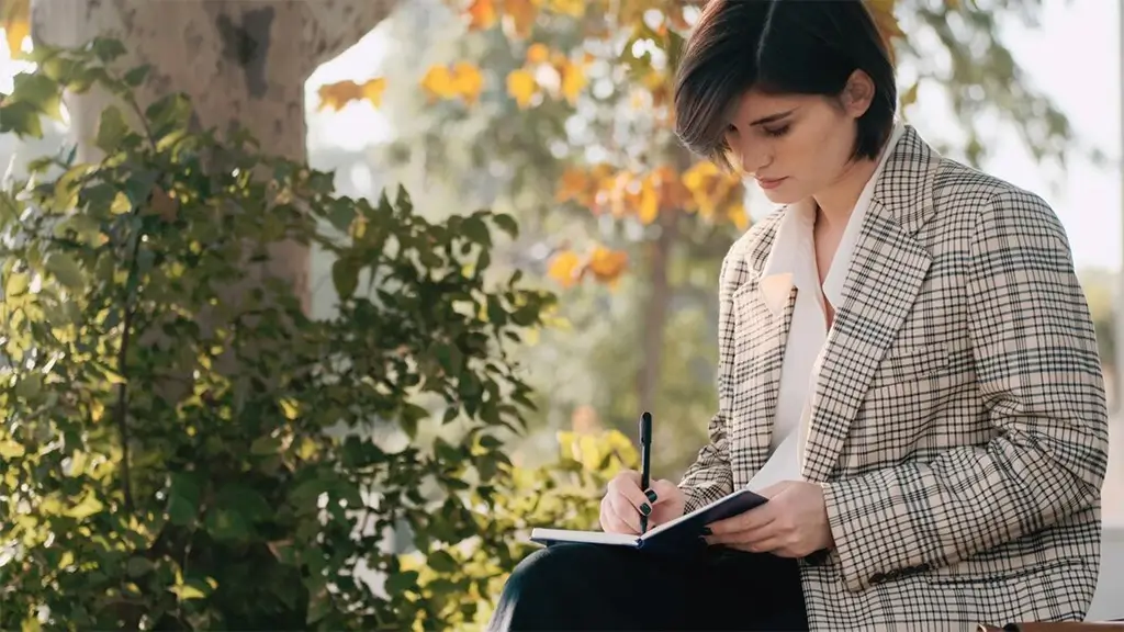 Woman in a plaid blazer writes in a notebook outdoors, seated among greenery on a sunny day.